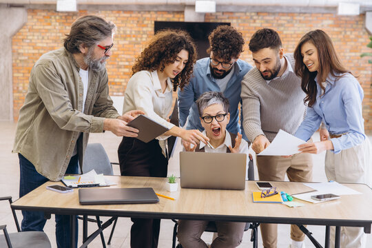 Stressed woman screaming at laptop during chaotic office meeting with team members bringing more work and creating pressure