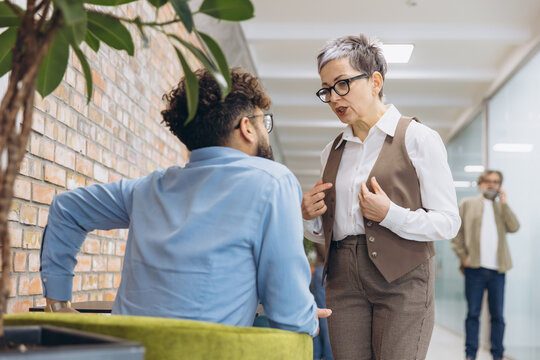 Mature female boss arguing with young male employee in office hallway, discussing corporate conflict