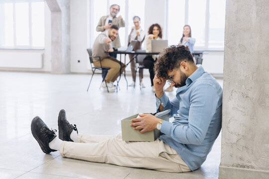 Sad man sitting on floor after being fired, colleagues in background pointing and cheering, experiencing humiliation and stress