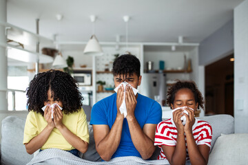 Sick African American family sitting on a sofa blowing their noses with tissues. Concept of seasonal flu, common cold, virus infection, healthcare at home, and family recovery during winter