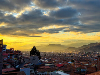 Obraz premium Sunset over Cusco city with traditional red rooftops, golden clouds, and snow-capped Andes mountains on the horizon, Peru