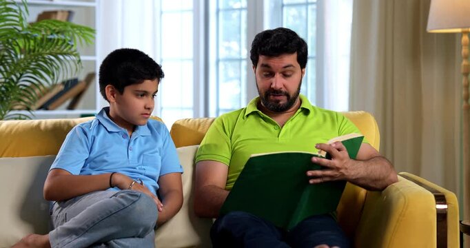 Indian asian father and son reading a book on sofa in modern home while dad explains school chapter and tells story to small kid sitting together in cozy living room family education bonding moment