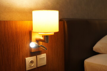 A modern bedside wall lamp mounted on a wooden panel, softly illuminating a cozy bedroom interior. The warm light creates a comfortable and relaxing atmosphere.