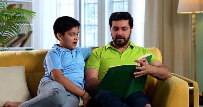 Indian asian father and son reading a book on sofa in modern home while dad explains school chapter and tells story to small kid sitting together in cozy living room family education bonding moment