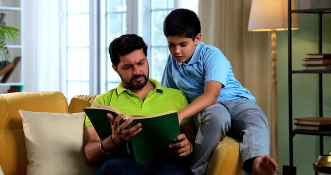 Indian asian father and son reading a book on sofa in modern home while dad explains school chapter and tells story to small kid sitting together in cozy living room family education bonding moment