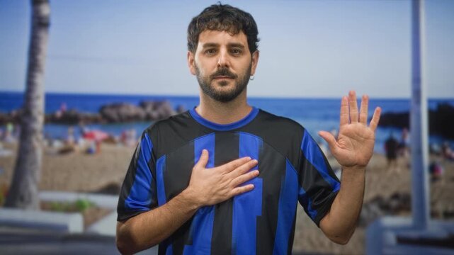 Young man with beard, hand on chest and left hand raised in pledge on a street by the beach; sincere.