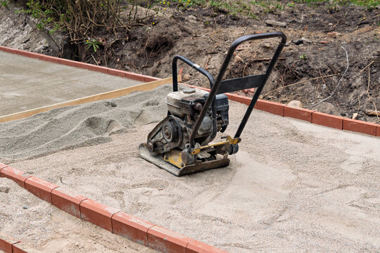Plate compactor rests on sand base inside curb as paving walkway is prepared during renovation at landscaping construction site.