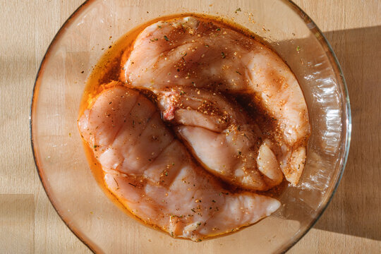 Raw chicken breasts marinate in spicy herb oil inside glass bowl on wooden board in warm sunlight, top view for home cooking prep.