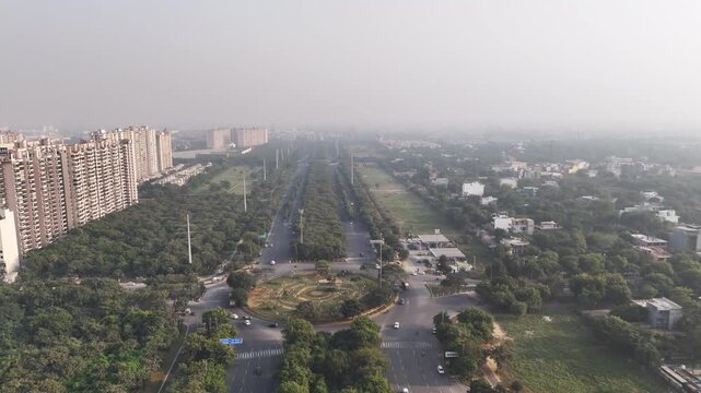 Aerial view of Pari Chowk, Greater Noida, showcasing the iconic roundabout, wide tree-lined avenues, and surrounding residential sectors under light haze, highlighting the city&rsquo;s planned urban layout.