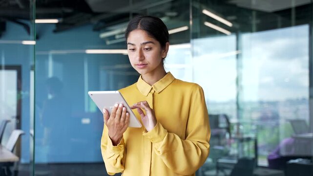 Smiling young businesswoman standing in modern office using digital tablet. Cheerful female employee browsing information, reading reports, or managing online tasks while staying productive at work.