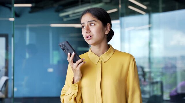 Upset young businesswoman standing in modern office talking on smartphone. Unhappy female employee feels disappointed after rejection or failed deal, reacting emotionally to bad news at work.