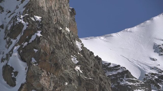 Panoramic snowy mountain range shows jagged rocky peaks, ridgeline contours, clear sky. Wide cinema vista reveals ice covered highland summits, crags, crest forms, azure backdrop.