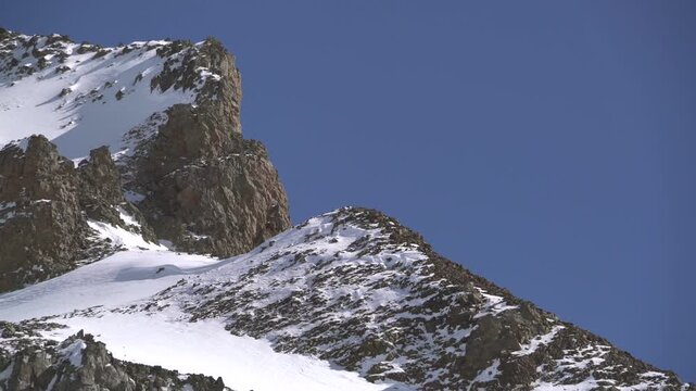 Panoramic snowy mountain range shows jagged rocky peaks, ridgeline contours, clear sky. Wide cinema vista reveals ice covered highland summits, crags, crest forms, azure backdrop.