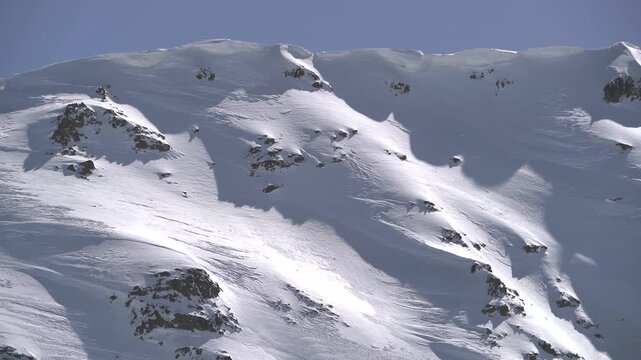 Panoramic snowy mountain range shows jagged rocky peaks, ridgeline contours, clear sky. Wide cinema vista reveals ice covered highland summits, crags, crest forms, azure backdrop.