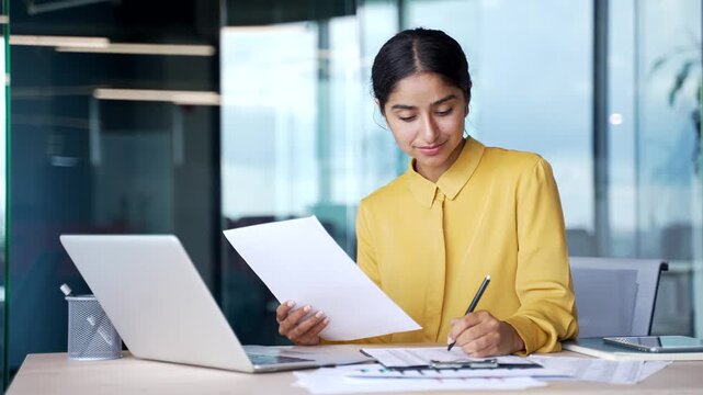 Busy businesswoman sitting at desk in office holding financial document and filling out form on table. Focused female accountant reviewing papers, writing notes, and doing bookkeeping or finance work.