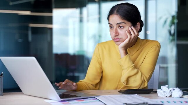 Young bored businesswoman sitting at desk in modern office feeling tired and unmotivated while working on laptop. Exhausted female employee struggling with fatigue, monotony, and lack of concentration