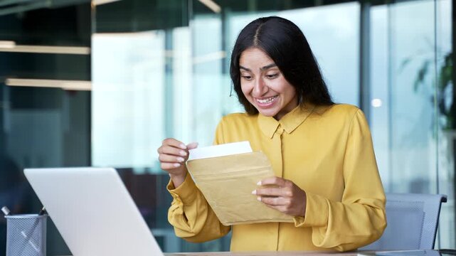 Happy young businesswoman sitting at desk in modern office reading letter with great news. Smiling cheerful female employee feeling excited and pleased after receiving positive message or document