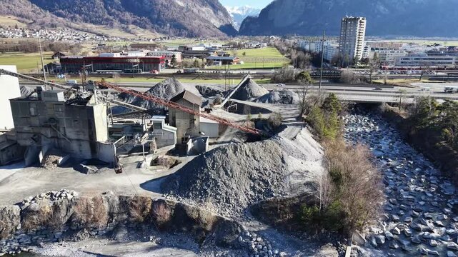 Aerial footage moving sideways across an industrial sand and gravel processing facility with visible aggregate piles and processing buildings. A highway with moving cars is visible in the background, 