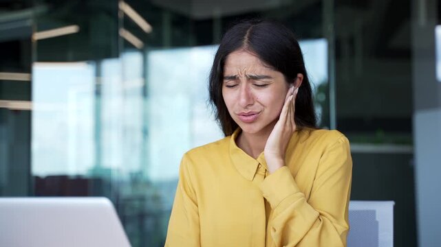 Close-up of young businesswoman touching her ear and wincing from pain in office. Female employee suffering from earache, feeling discomfort and irritation caused by ear infection or inflammation.