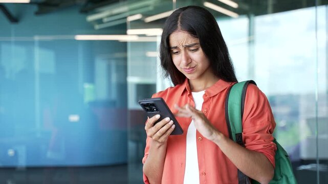 Frustrated young female student standing in modern university building having trouble using smartphone. Confused woman with backpack looking at phone, experiencing technical problem or app error.