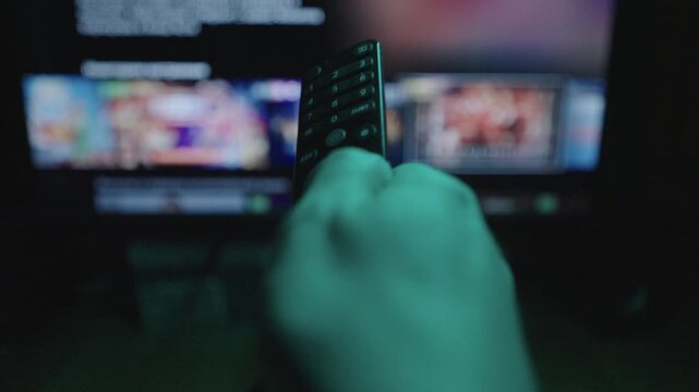 Close-up of hand pressing buttons on tv remote, navigating video streaming platform displaying various movie and show options on television screen, enjoying cozy night of home entertainment.