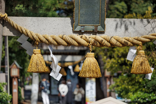 Sacred shimenawa straw rope and zigzagged shide paper streamers hang from sunlit concrete torii gate, marking boundary to sacred space of small neighborhood Shinto shine.