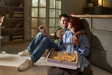 Young couple sitting on floor eating pizza together, smiling and looking at each other, open pizza...