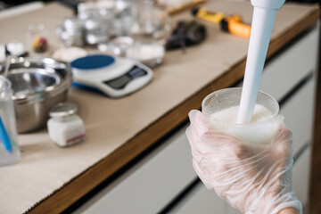 Person using immersion blender to mix liquid base in glass jar on digital scale. cosmetic emulsifying, professional cream blending, skincare laboratory tools, making lotion process.