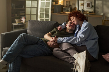 Fototapeta premium Caucasian young adult couple relaxing on sofa, woman with curly red hair leaning head on man's shoulder, both holding hands and smiling, cozy home interior in background