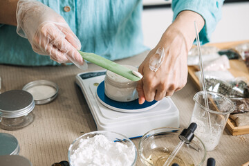 Person in gloves applies cream from spatula to hand while measuring jar on digital scale. Dermatological testing, plant-based lotions, organic product design, eco-conscious beauty.