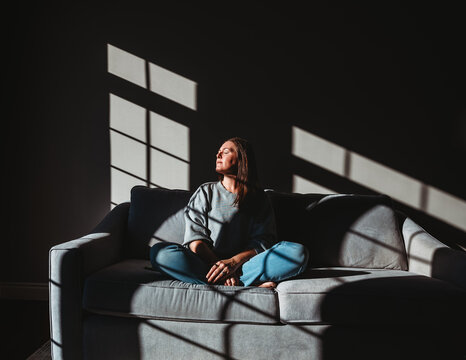Woman sitting on sofa in dark room in patch of sunlight from window.
