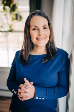 Smiling woman in blue top standing by window with folded hands