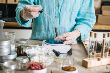 Gloved hands use glass dropper to add liquid to botanical ingredients in test tube on digital scale. Liquid boosters, floral droplets, herbal vials, custom blends.