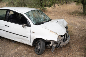 Crashed white sedan beside dry oak trees, crumpled front end, shattered windshield, detached bumper, flat front wheel, dirtstrewn ground,