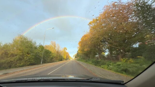 View from inside moving car driving along country road toward bright rainbow arch in autumn landscape. Natural light, colorful sky and road perspective create concept of hope, journey, optimism, new