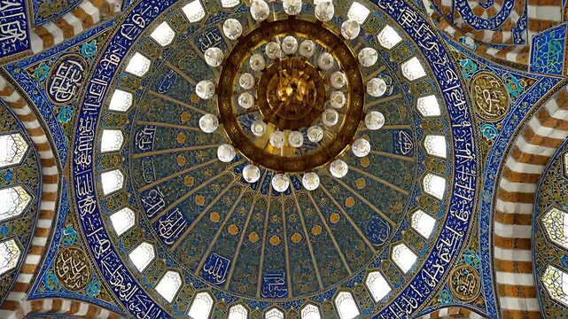 Intricate mosaic tilework and ornate calligraphy adorn the magnificent interior dome ceiling of a traditional Islamic mosque featuring a large central chandelier.
