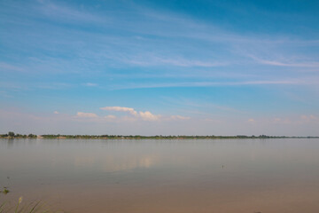 Vast tranquil river landscape reflecting the soft blue sky over a distant, low tropical village horizon.