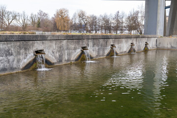Naklejka premium Flowing water from several rectangular spouts enters a green water basin near a bridge during winter day