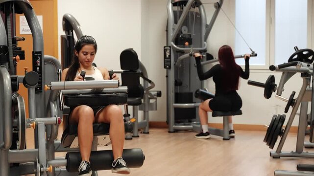 Young woman working out on a leg curl machine at the gym while a female personal trainer approaches to give her instructions and guidance
