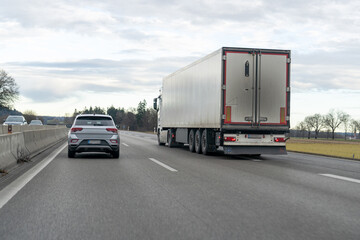 Obraz premium Silver SUV driving behind large semi-trailer truck on a multi-lane highway on an overcast day
