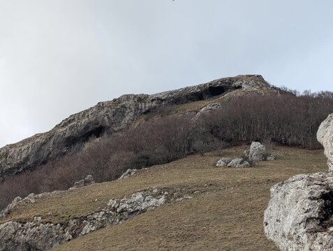 Pendio montano roccioso con erba secca, rocce affioranti e alberi spogli sotto un cielo nuvoloso