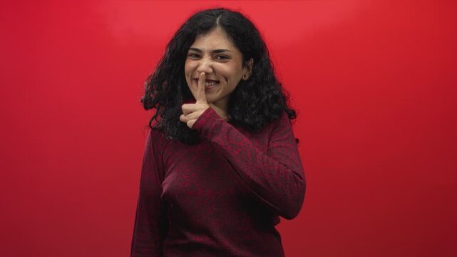 Young arab brunette woman finger to lips for silence in red studio with curly hair and dimpled smile; playful mischief.