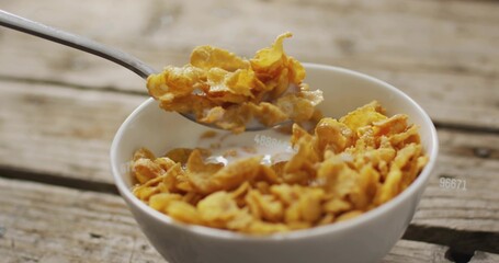Naklejka premium Scooping metal spoon pulling golden corn flakes from white china bowl on wood planks, milk clinging