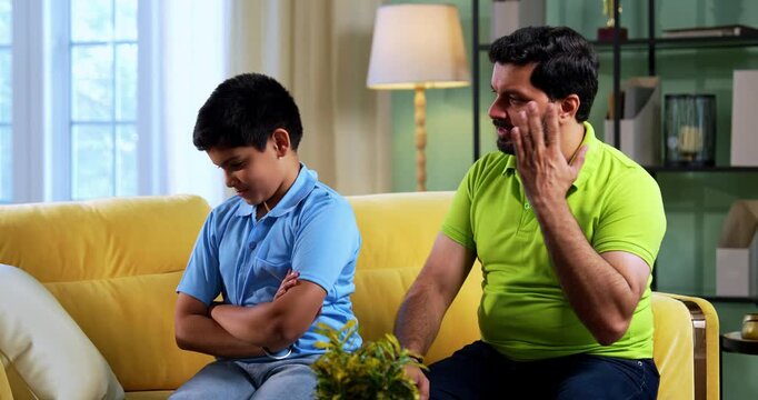 Indian asian young father scolding son while sitting on sofa in modern home as upset child turns back silently, showing emotional parenting moment and family tension inside living room