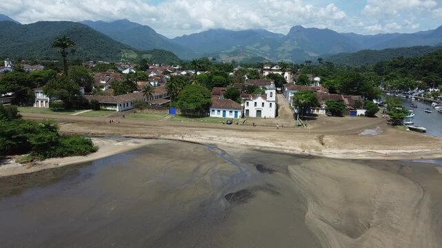 Drone shot flying toward Paraty town near the river mouth, featuring Igreja Nossa Senhora das Dores on Prainha do Forte beach. Includes surrounding mountains and Atlantic forest in the background.
