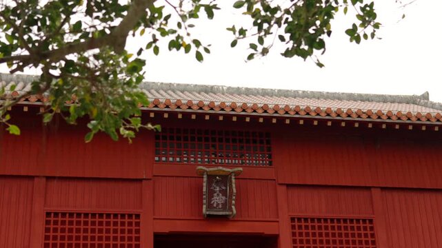 Static close-up shot of the vibrant red wooden facade and tiled roof of Shuri Castle&rsquo;s main gate, partially framed by green foliage against a bright overcast sky. Features a central plaque with ornate