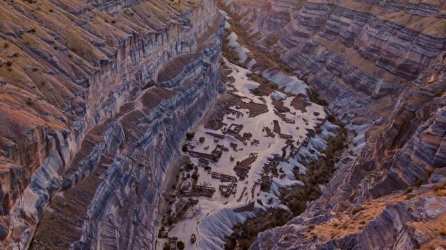Golden sunset above steep layered canyon walls revealing ancient ruined settlement on pale sandy valley floor, concept of a lost civilization and the grandeur of geological epochs