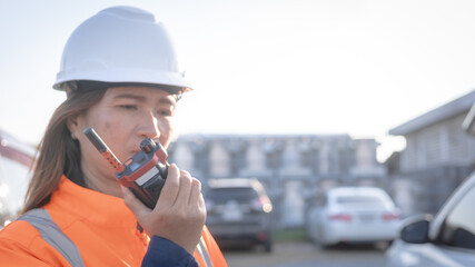 A construction worker stands at a site and uses a radio. This takes place during the day in a suburban area with buildings and vehicles in the background © Happy Photo