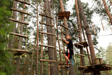 young toddler boy climbing over obstacle with rope in adventure nature activity park. continuous belay system on him. harness and safety line