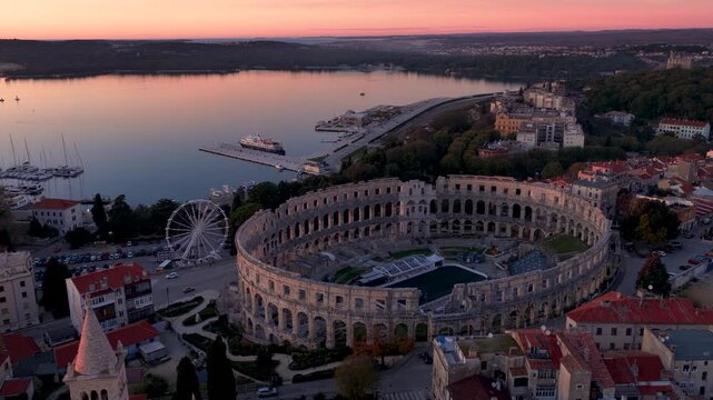Aerial view of the ancient Pula Arena, a grand amphitheater, juxtaposed with the serene harbor under a soft, pastel sky, Pula, Istria County, Croatia.